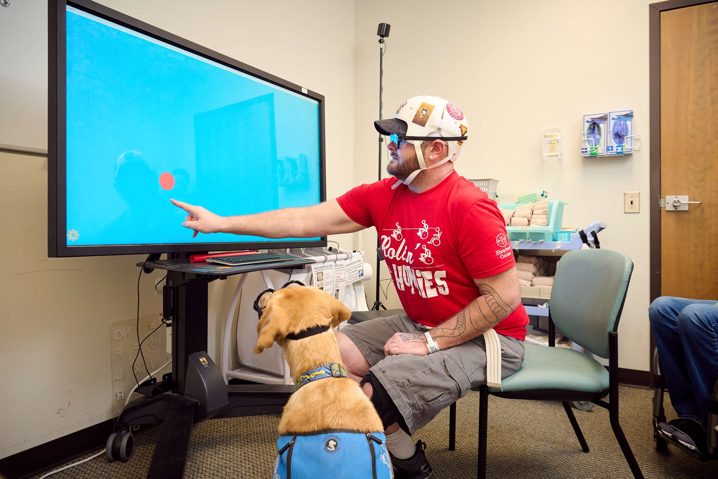 A person wearing a white helmet and red shirt sits in a chair, pointing at a large screen with a red dot. A service dog, wearing a blue vest, sits attentively at their feet. Medical equipment is visible in the background.