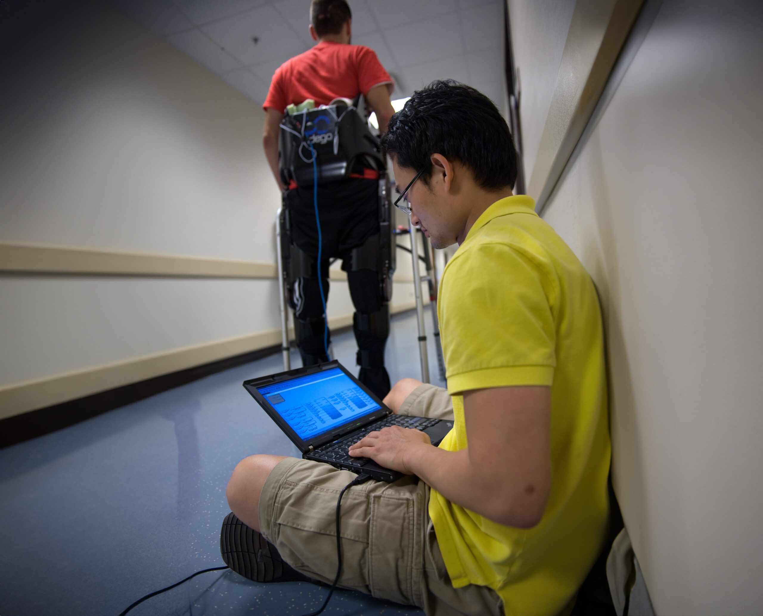 A person in a yellow shirt and shorts sits on the floor in a hallway, working on a laptop. In the background, another person wearing a red shirt and using a walker equipped with a robotic exoskeleton walks away from the camera.