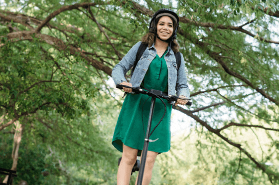 A woman wearing a green dress and a denim jacket rides a scooter under a canopy of lush green trees. She is smiling and wearing a helmet. The background is sunlit, creating a serene outdoor setting.