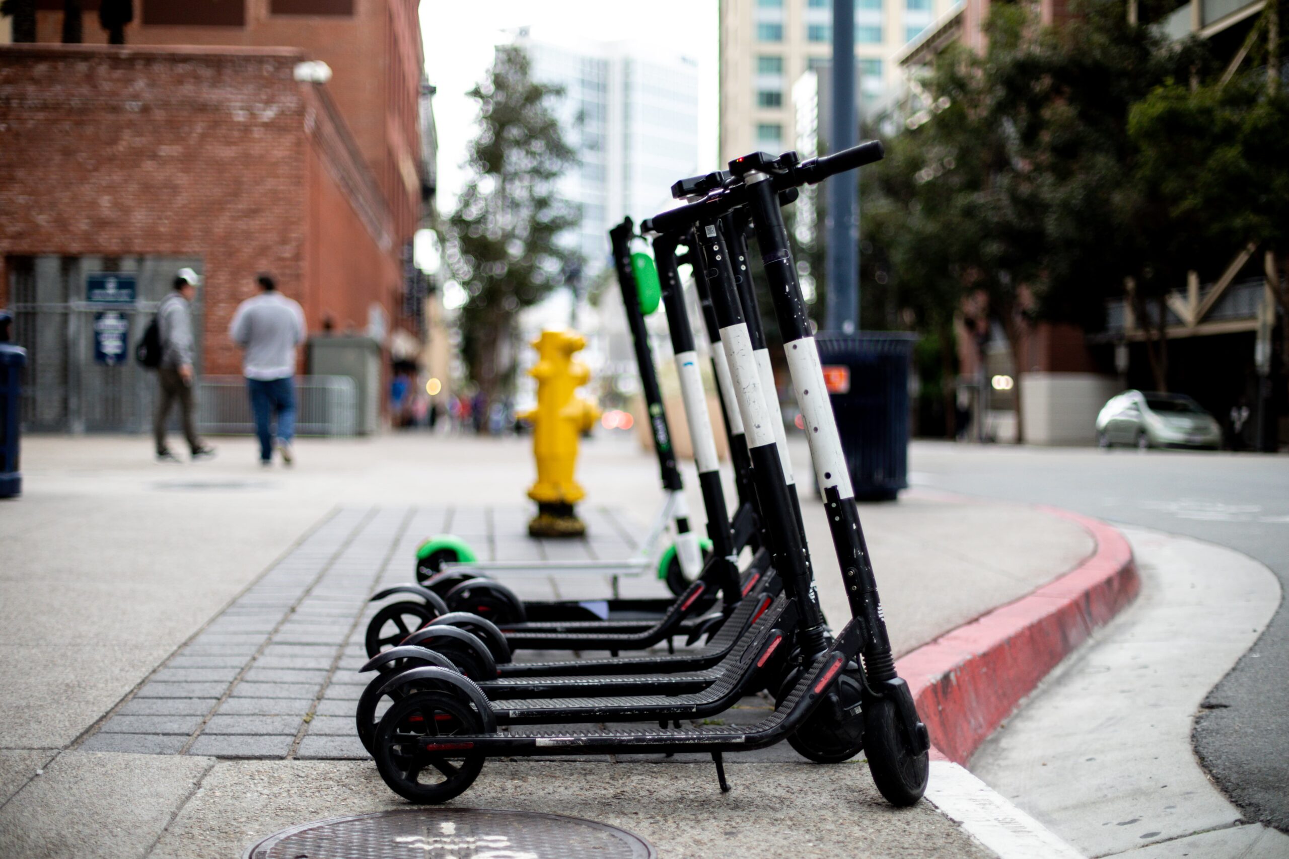 A row of electric scooters is parked on a city sidewalk near a street corner. A yellow fire hydrant is visible in the background, along with a few pedestrians and tall buildings.