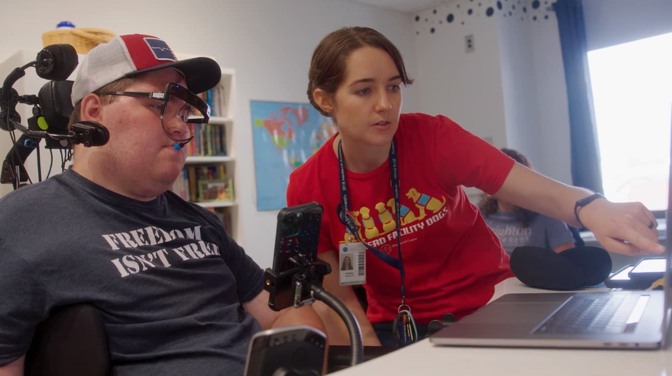 A person in a wheelchair uses a mouth-operated device to interact with a laptop, while a woman in a red shirt points at the screen, assisting. They are in a room with a world map on the wall and bookshelves in the background.