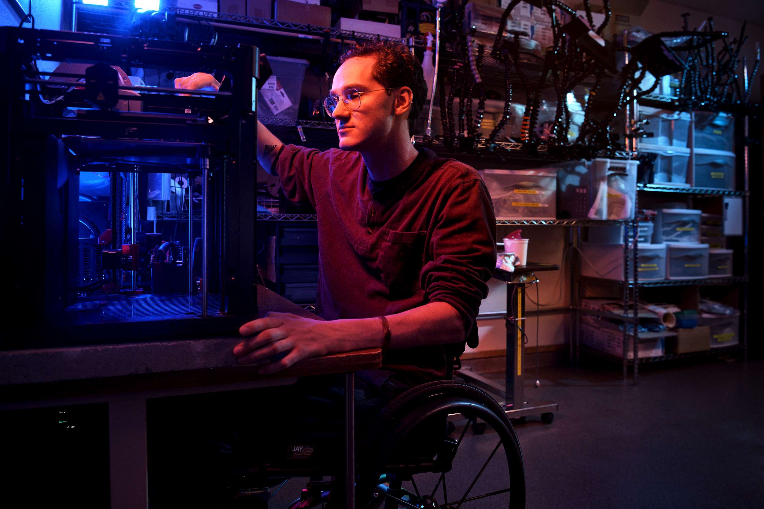 A person in a wheelchair is adjusting a 3D printer in a dimly lit workshop. They are wearing glasses and a dark long-sleeve shirt. The room is filled with shelves of tools and parts, illuminated by blue and purple lighting.
