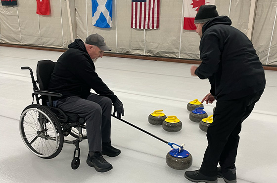 A person in a wheelchair and another person are curling on an ice rink. Several stones with yellow and blue handles are on the ice, and flags of various countries hang in the background.