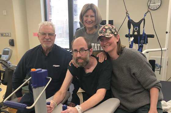 A group of four people in a physical therapy setting. A man sits on an exercise machine, smiling. Two women and another man stand around him, all smiling. There's exercise equipment and a clock on the wall in the background.