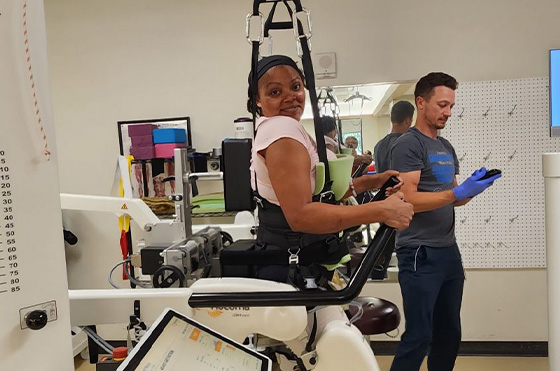 A woman smiles while using an exoskeleton machine in a rehabilitation setting. She is supported by a harness. A man in the background is checking a device. Various medical equipment is visible.