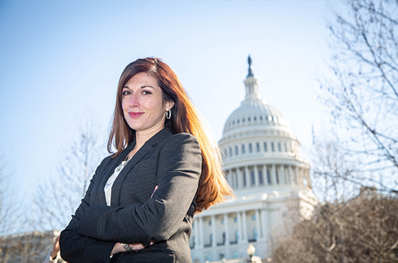 Woman with long brown hair stands confidently in front of the U.S. Capitol building, wearing a dark blazer and smiling at the camera. The sky is clear and blue, with some bare tree branches visible.