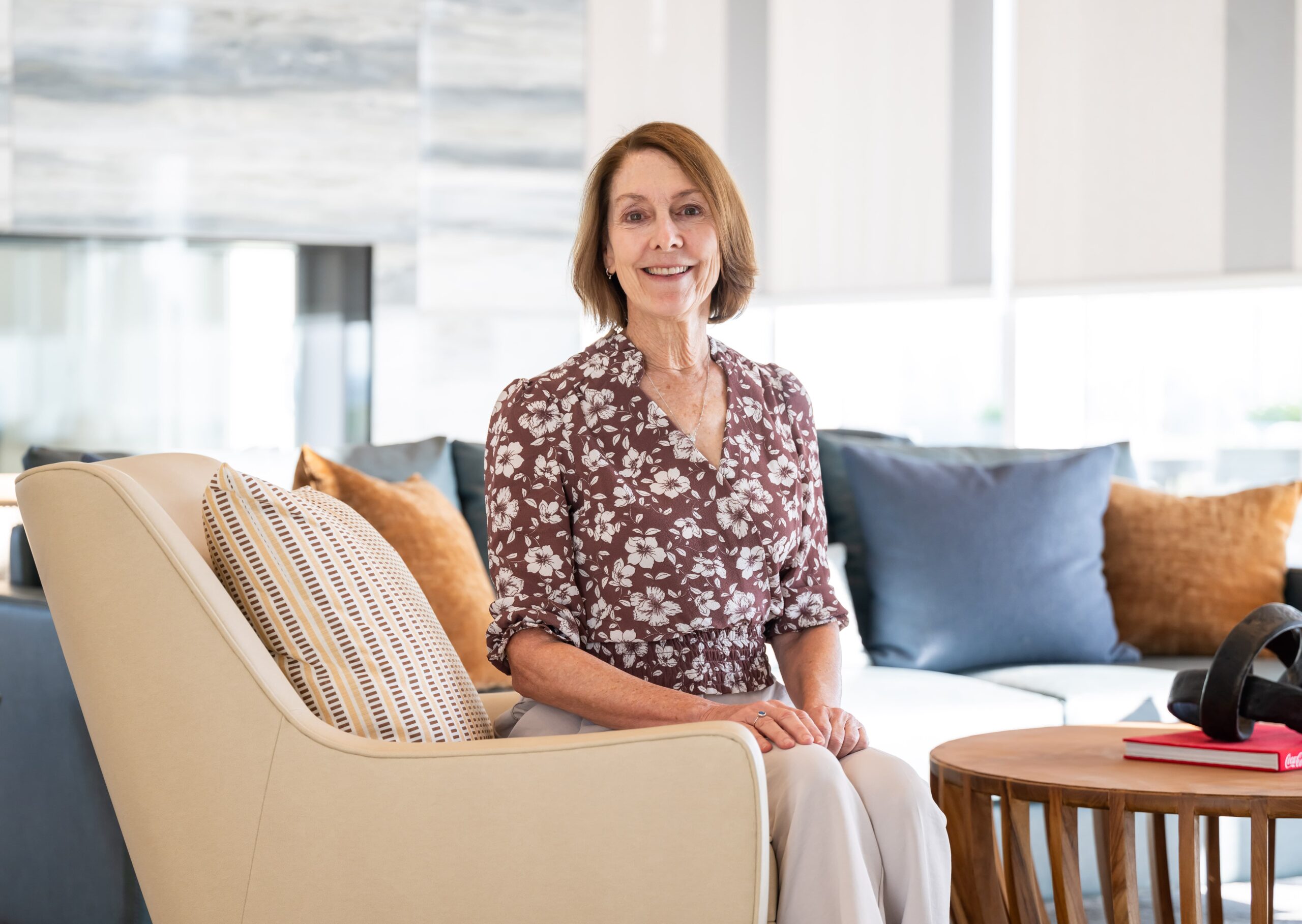 Woman wearing a floral blouse sits on a beige chair in a bright living room with white and brown cushions. She is smiling and has her hands resting on her lap. A wooden table with a book and headphones is visible in the foreground.