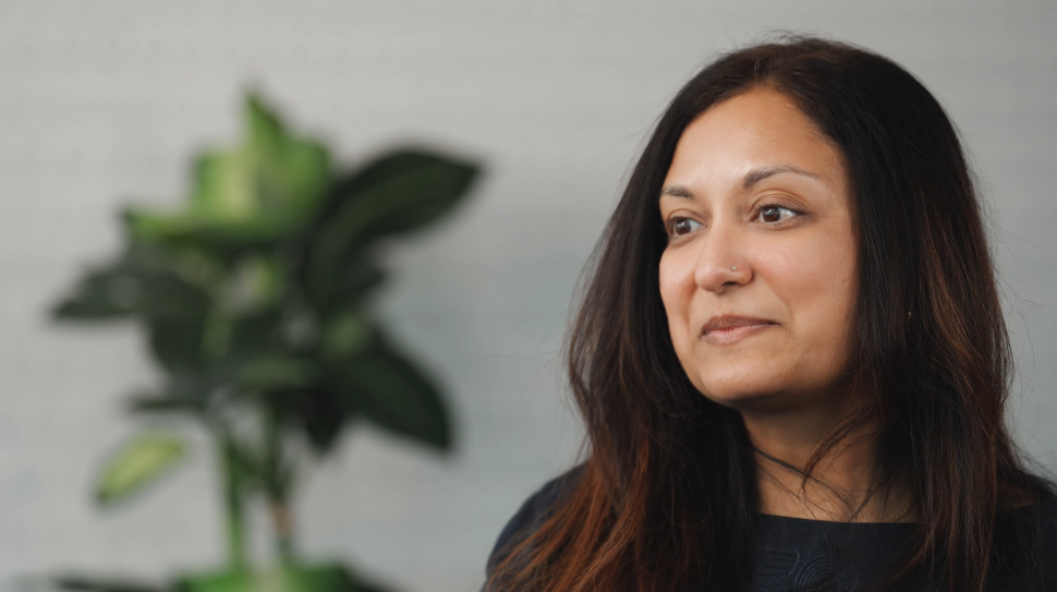 A woman with long dark hair and a dark top is smiling softly, looking slightly to the side. A green leafy plant is blurred in the background against a light wall.