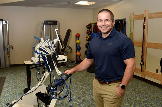 A person in a blue shirt stands smiling next to exercise equipment in a fitness room at the renowned Shepherd Center, known for its awards and recognition. The room includes various gym machines, colorful exercise balls, and resistance bands mounted on the wall.
