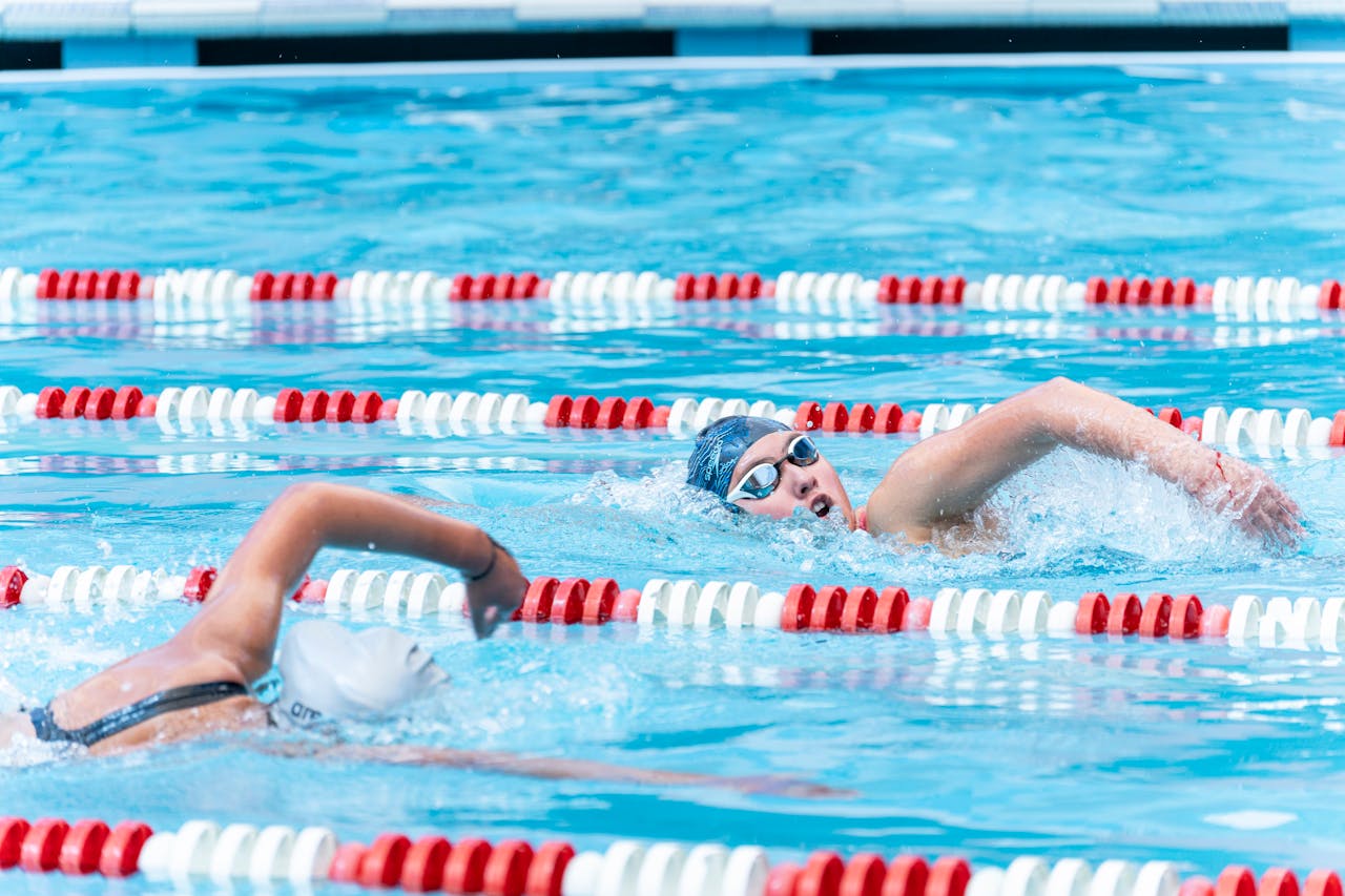 Two swimmers wearing swim caps and goggles compete in a pool, performing freestyle strokes in adjacent lanes marked with red and white lane dividers.