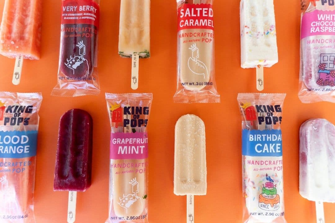 An assortment of colorful King of Pops popsicles, both unwrapped and in clear packaging, arranged neatly on an orange background. Flavors include Very Berry, Salted Caramel, Grapefruit Mint, Blood Orange, and Birthday Cake.