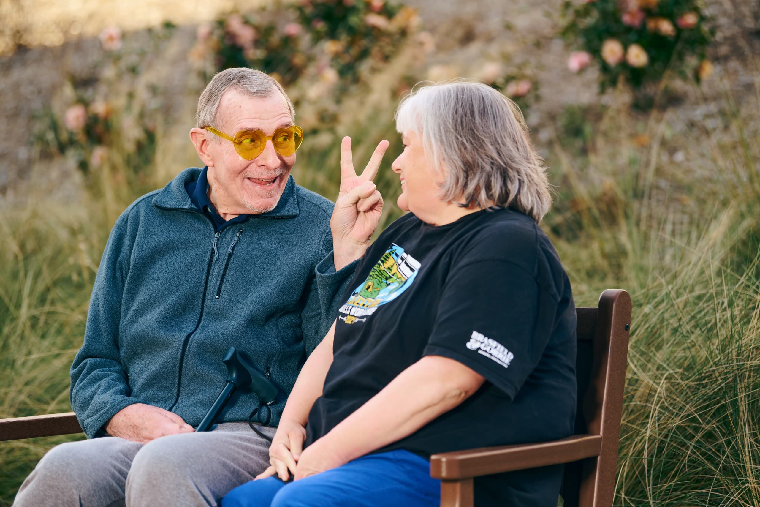 An older man wearing yellow glasses playfully makes a peace sign behind an older woman’s head as they sit together on a bench outdoors, both smiling and enjoying each other's company. Shrubs and flowers are in the background.