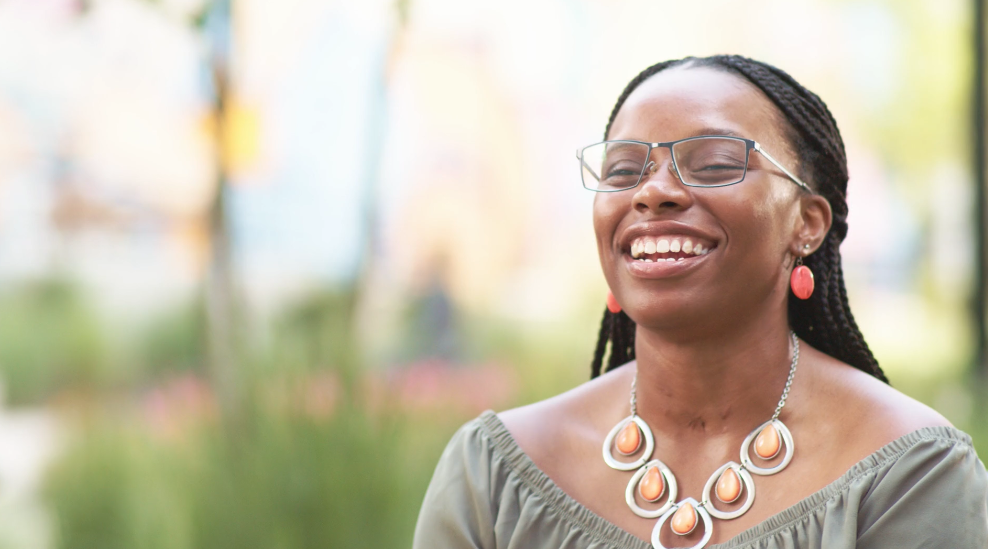 A woman with braided hair and glasses is smiling brightly outdoors. She is wearing a gray off-the-shoulder top, orange earrings, and a matching statement necklace. The background is blurred greenery.