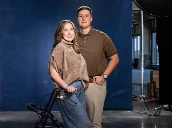 A smiling woman stands next to a man in front of a blue backdrop. The woman holds onto a walker, wearing a brown top and blue pants. The man stands behind her, wearing a brown shirt and beige pants. Studio lights are visible in the background.