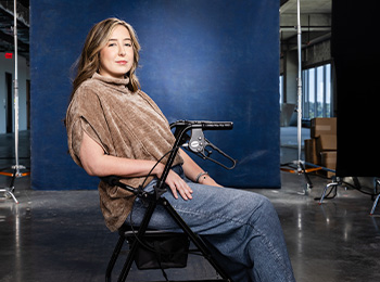 A woman with light brown hair sits confidently on a rolling walker in front of a blue backdrop in a photography studio, wearing a tan top and blue jeans. Natural light streams through large windows in the background.