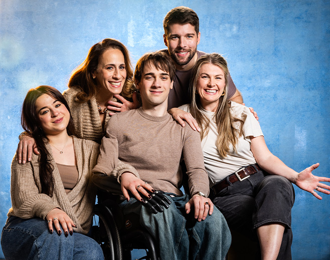 Five young adults, including a person in a wheelchair, sit and stand closely together, smiling against a blue background. They appear happy and supportive, dressed casually in neutral colors.