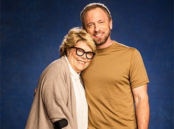 A smiling woman with short blond hair, glasses, and a beige cardigan stands closely with a smiling man in a tan shirt against a blue background. They appear happy and comfortable together.