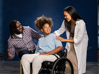 A smiling girl in a wheelchair sits between a man and a woman, who both lean in close and smile warmly at her against a dark blue background.