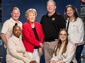 Six people pose for a group photo in front of a blue backdrop. Three are standing in the back, while three are seated in front. They are all smiling and wearing business casual clothing.